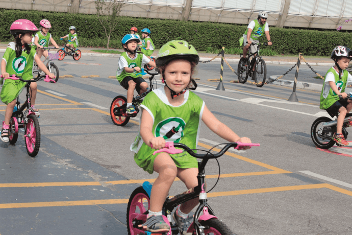 Several children drive in bikes at the schools car parks. They wear security wests and helmets.