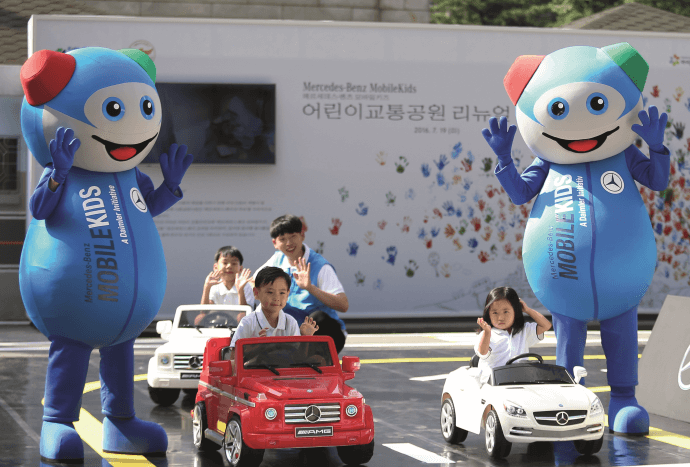 Four children drive pedal cars on a parcours. Two people in Moki-costumes are right next to the driving lane.